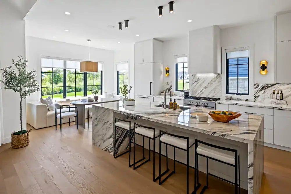 Modern kitchen with white cabinets, marble countertops, and island with barstools. Large windows let in natural light, and a dining area with cushioned seating is visible. Wooden floors and minimalist decor complete the space.