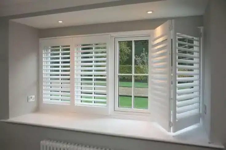 A white-framed window with plantation shutters; two shutters are closed while one on the right is open, revealing a green outdoor landscape. The window sits in a recessed wall with a wide windowsill.